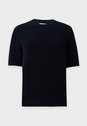 Un mannequin se tient devant un fond blanc uni, vêtu d’un haut noir, d’un jean bleu et de chaussures noires, avec un sac noir porté à l’épaule.