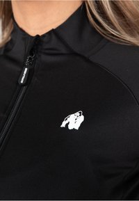 Chaqueta negra con cremallera hecha de tela suave, que presenta un logo de gorila blanco en el lado izquierdo del pecho, con un cuello alto y un diseño elegante.