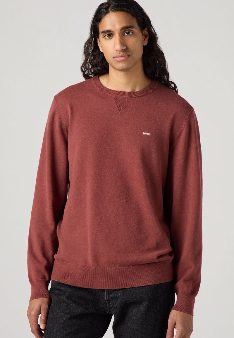 Pull maroon en tricot doux avec un col rond et des poignets côtelés. Présente un petit logo sur la poitrine et une coupe classique.