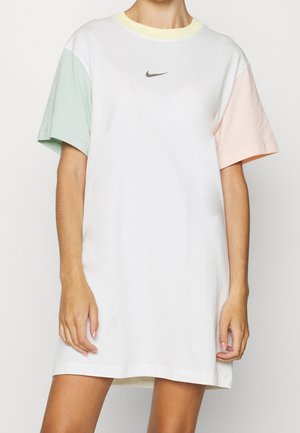 Biała oversize sukienka T-shirt marki Nike z zielonym lewym rękawem, różowym prawym rękawem i żółtym dekoltem noszona przez stojącą osobę.