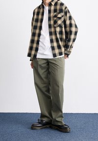 Svart och beige rutigt skjorta med knappar, vit t-shirt med rund hals, olivgröna byxor och svarta skor med kraftig sula.