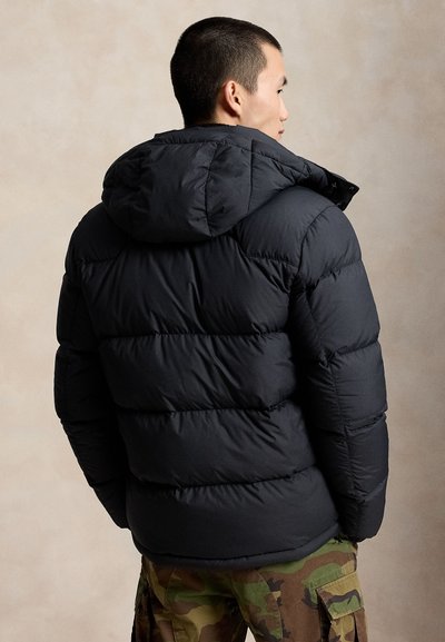 Man wearing black puffer jacket with hood and camouflage cargo pants, facing sideways against a plain beige background.