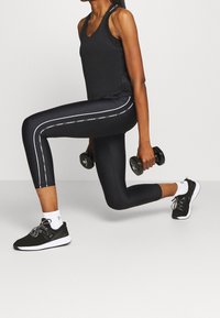 Svart linne och leggings med vita sidostråkar, texturerad tyg, knälång. Svarta träningsskor och hantlar hålls i varje hand.