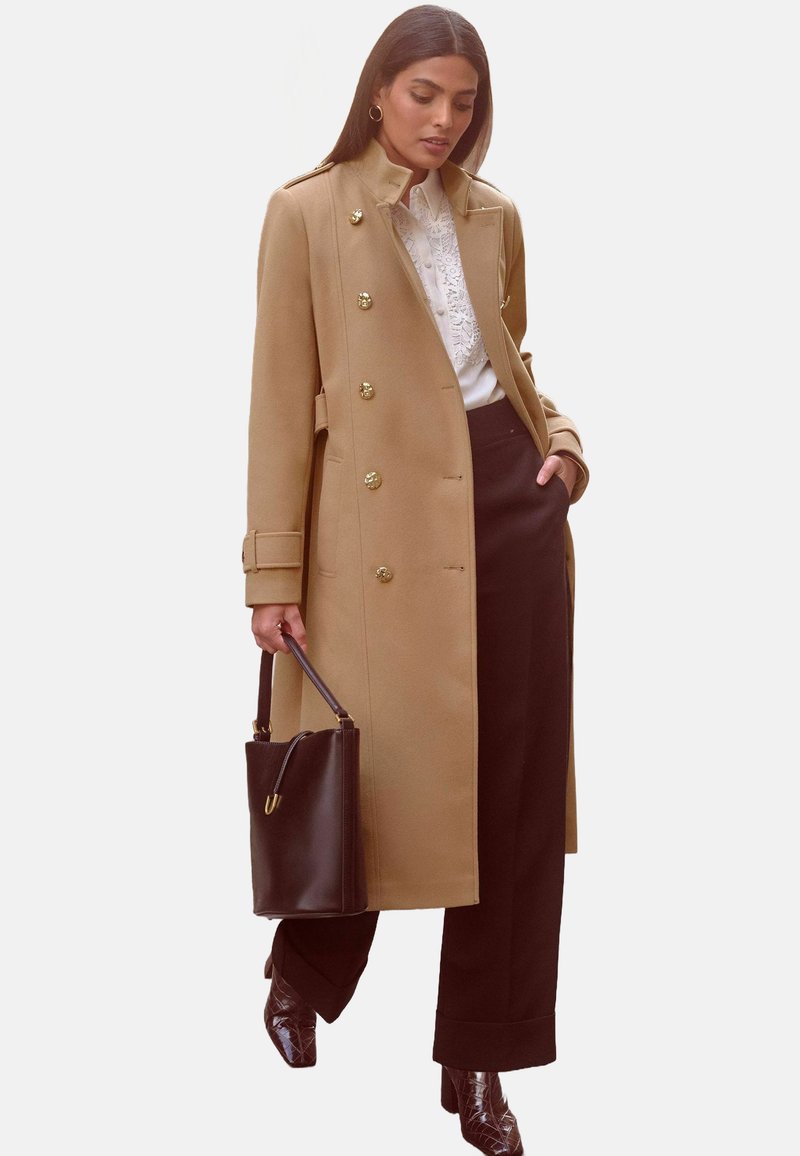 Beige Wollmischungsjacke mit goldenen Knöpfen, hohem Kragen und taillierter Passform. Kombiniert mit schwarzen Hosen und einer dunklen Handtasche. Glänzende braune Stiefel mit Absatz.
