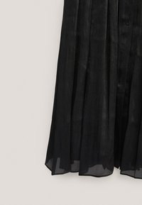 Jupe plissée semi-transparente noire ou ourlet de robe avec détail bouton, suspendue sur un fond beige clair.