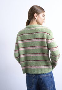 Νεαρή γυναίκα με ανοιχτό καστανά μαλλιά μαζεμένα σε αλογοουρά, φορώντας πράσινο πλεκτό πουλόβερ με ρίγες κόκκινες και ροζ, συνδυασμένο με μπλε τζιν, κοιτάει προς τα πλάγια.