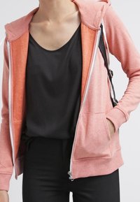 Mujer con una sudadera con cremallera color coral sobre una camiseta sin mangas negra, con una mano en el bolsillo de la sudadera y una correa de mochila negra en el hombro.
