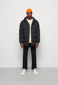 Giacca nera imbottita con cappuccio, caratterizzata da bottoni frontali e dettagli di tasche con zip. Abbbinata a una camicia beige, pantaloni neri e sneakers bianche.