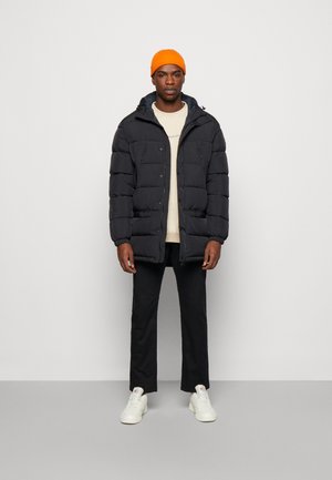 Giacca nera imbottita con cappuccio, caratterizzata da bottoni frontali e dettagli di tasche con zip. Abbbinata a una camicia beige, pantaloni neri e sneakers bianche.