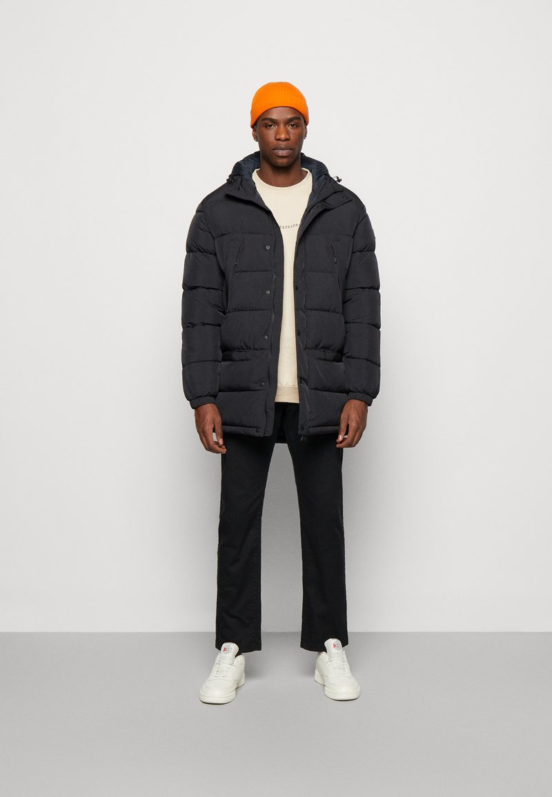 Giacca nera imbottita con cappuccio, caratterizzata da bottoni frontali e dettagli di tasche con zip. Abbbinata a una camicia beige, pantaloni neri e sneakers bianche.