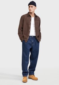 Chemise en velours côtelé marron, t-shirt blanc, jeans bleu baggy et chaussures en daim beige, associés à un bonnet bleu marine. Tenue décontractée avec une coupe ample.
