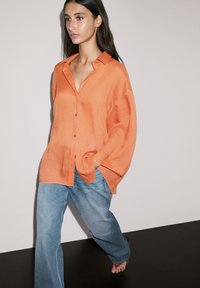 Oranje linnen shirt met lange mouwen, een kraag en knoopsluiting, gecombineerd met lichtblauwe wijde denim broek. Effen achtergrond.