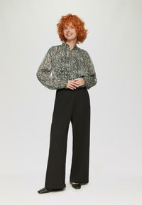 Doorzichtig groen blouse met patronen en pofmouwen, gecombineerd met zwarte wijde broek en zwarte platte schoenen, op een effen achtergrond.