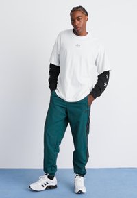 Hvit t-skjorte med en liten logo, svart langermet skjorte, teal joggebukser og hvite joggesko med svarte detaljer. Enkel og sporty design.