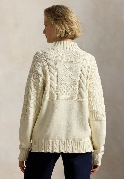 Γυναίκα που φοράει ένα κρεμ πουλόβερ με πλεξούδες και σχέδιο με σταυρωτά μπέιζμπολ ρόπαλα στην πλάτη, συνδυασμένο με σκούρο παντελόνι, με την πλάτη γυρισμένη στην κάμερα.