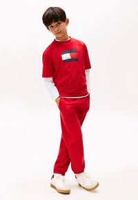 Chlapec stojí s rukami v kapsách, oblečený v červených teplákoch, červenom tričku s bielymi a námořníckymi pruhmi, bielej košeli s dlhým rukávom a bielych teniskách.