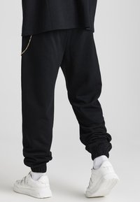 Svarta joggarbyxor i mjukt tyg, med en tapered passform, elastiska muddar och en guldkedjedetalj på sidofickan. Matcher med vita sneakers.