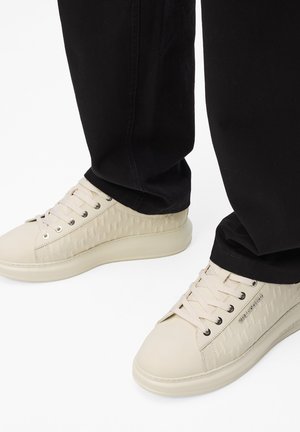 Sneakers beige in pelle con pannelli laterali testurizzati e lacci rotondi. Punta arrotondata e suola a profilo basso. Indossati con pantaloni neri.
