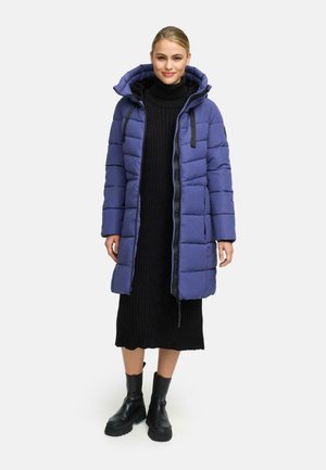 Eine Frau steht in einem langen blauen gepolsterten Mantel über einem schwarzen gerippten Kleid und schwarzen klobigen Stiefeln vor einem schlichten hellen Hintergrund.