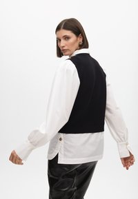 Schwarze Strickweste über einem weißen Hemd mit Knöpfen, die goldene Akzente und ein geschichtetes Design aufweist. Das Model wird von hinten gesehen.