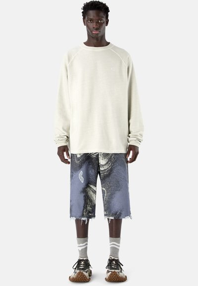 Modèle masculin portant un sweat-shirt crème surdimensionné, un short mi-cuisses à motifs bleus et noirs, des chaussettes grises et des baskets marron, debout sur fond blanc.