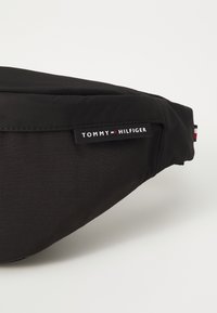 Svart nylon midjeveske med en glatt tekstur, som har et fremtredende hvitt "TOMMY HILFIGER"-merke og røde, hvite og blå detaljer.