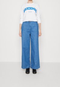 Weißes langärmeliges, cropped Shirt mit blauem "Abercrombie New York"-Text, kombiniert mit lockeren blauen Denim-weit geschnittenen Hosen und schwarzen Clogs.