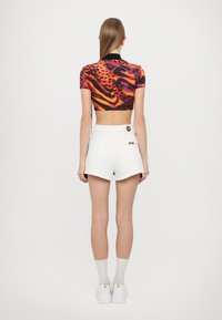 Top corto en negro, naranja y púrpura con estampado de tigre; combinado con pantalones cortos blancos que tienen un botón y detalles decorativos de metal.