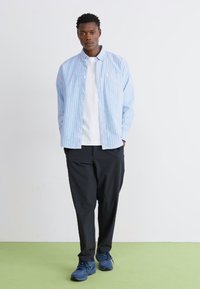 Lyseblå stribet skjorte med knapper over en hvid t-shirt, parret med løse sorte bukser og blå sneakers. Glat stof og afslappet pasform.