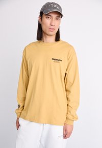 T-shirt en coton à manches longues, jaune moutarde, avec un logo imprimé noir sur la poitrine. Associé à un pantalon blanc, porté avec une casquette grise.