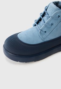 Botte en suède bleu clair avec une semelle en caoutchouc texturé bleu foncé et un capuchon de pied, dotée de lacets bleus passant par des œillets en métal.