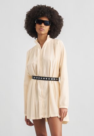 Femme aux cheveux bouclés volumineux portant des lunettes de soleil noires, une mini-robe plissée beige à manches longues et une ceinture noire cloutée.