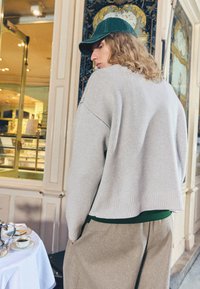 Svetro s pletenjem v svetlo sivi barvi, z odprtim krojem in rebrastimi manšetami ter robom. Kombiniran z olivno zeleno bluzo in bež širokimi hlačami.