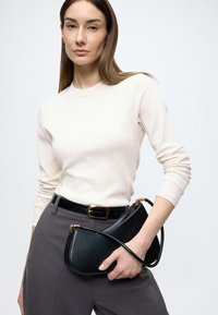 Schwarze Lederhandtasche mit geschwungener Form und goldenen Beschlägen, gehalten vor einem cremefarbenen, gerippten Oberteil und grauen Hosen, akzentuiert durch einen schwarzen Gürtel.