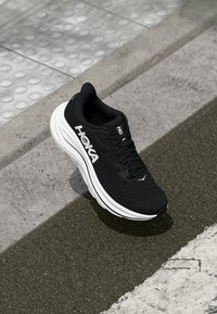 Chaussure de sport noire avec des caractéristiques comprenant une tige en maille, une semelle intermédiaire blanche épaisse et des lacets noirs. Logo bien en vue sur le côté.