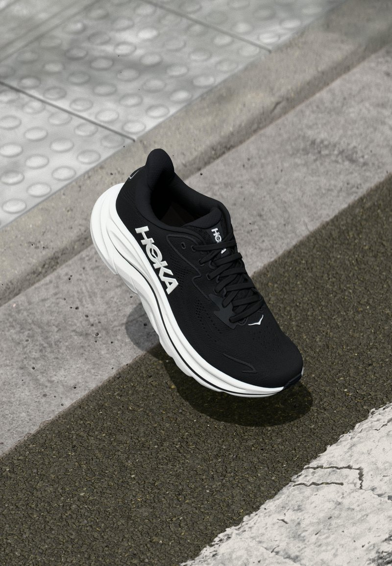 Chaussure de sport noire avec des caractéristiques comprenant une tige en maille, une semelle intermédiaire blanche épaisse et des lacets noirs. Logo bien en vue sur le côté.
