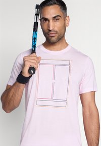 Ροζ βαμβακερό μπλουζάκι με πολύχρωμο κεντητό γραφικό σχέδιο και κεντρικό λογότυπο Nike. Το μοντέλο κρατά μπλε και μαύρη ρακέτα τένις.