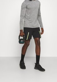 Graues langärmliges Sportshirt mit strukturiertem Finish, kombiniert mit schwarzen Shorts mit neongrünen Akzenten. Hält eine schwarze Kettlebell.