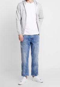 Henkilö, joka on pukeutunut vaaleanharmaaseen vetoketjulliseen huppariin valkoisen T-paidan päällä, vaaleansinisisiin kulutettuihin farkkuihin ja valkoisiin lenkkareihin, seisoo tasaisella taustalla.