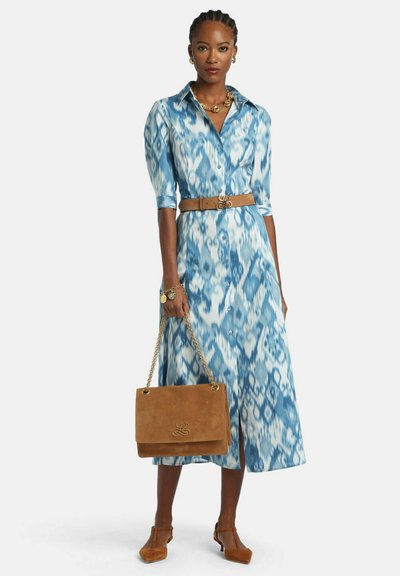 Žena v midi šatách s modro-bielym vzorom, s opaskom v hnedej farbe, drží hnedú kabelku so zlatými reťazovými remienkami a má na sebe dokonale zladené hnedé lodičky.