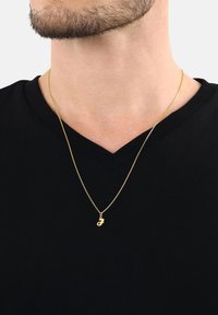 Collar de oro con un pequeño colgante, llevado sobre una camiseta negra de cuello en V; la cadena de oro es delgada y delicada, mostrando un acabado suave.