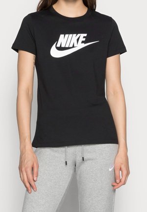 Osoba ubrana w czarną koszulkę Nike z białym logo oraz jasnoszare dresowe spodnie Nike, stojąca z rękami swobodnie opuszczonymi wzdłuż ciała.