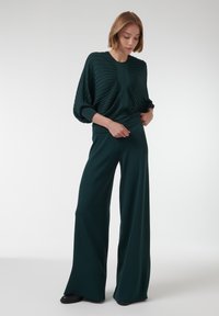 Donkergroene gebreide outfit met een geplooide, oversized top en wijde broek, beide met een gladde textuur en een relaxte pasvorm. Zwarte schoenen maken de look compleet.