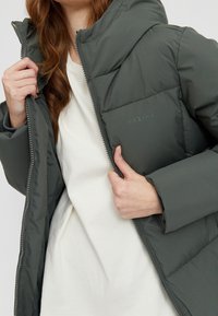 Veste matelassée vert foncé avec capuche, fermeture éclair à l'avant et poignets élastiques. Présente un design matelassé et un logo de marque subtil sur la poitrine.