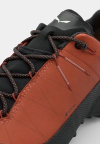Obuv GORE-TEX s oranžovým syntetickým zvrškom s textúrovaným vzorom, čiernou sieťovinou a kontrastnými tmavými šnúrkami. Pevná gumová podrážka.