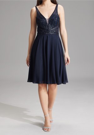 Femme portant une robe bleu marine sans manches mi-longue avec un corsage à sequins et une jupe fluide, associée à des talons argentés à bride à la cheville.