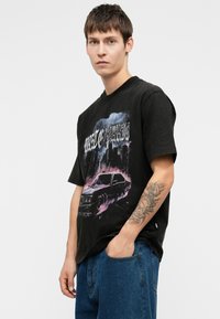 Mlad muž s kratkom tamnom kosom koji nosi crnu grafičku majicu s dizajnom automobila i plave traperice, stoji naslonjen na bijelu pozadinu.