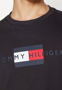 Close-up of a person wearing a dark shirt with a rectangular Tommy Hilfiger logo featuring navy, white, and red color blocks on the chest.