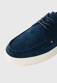 Ναυτικές σουέτ παπούτσια με στρογγυλή μύτη, λευκή καουτσούκ σόλα και γκρι κορδόνια. Διαθέτει ραφή κατά μήκος της άκρης και ένα μικρό κόκκινο, λευκό και μπλε λογότυπο.