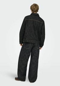 Melna džinsa jaka un platām kāju bikses, uzsverot neapstrādātu faktūru, kontrastējošas dzeltenas šuves un aizmugures kabatas ar klasisku dizainu.